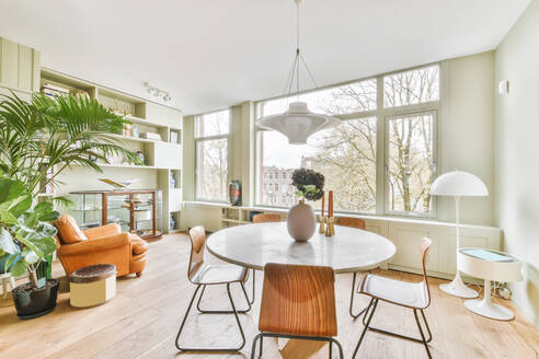 Round table with flowers and candlesticks placed in stylish light living room with armchair and green potted plants in apartment with large windows - ADSF35097