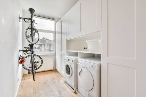 Moderne weiße elektrische Waschmaschinen und Trockner in der Nähe von weißen Schränken und Körben in einer hellen Waschküche mit Fenster und Fahrrad - ADSF35102