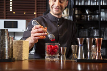 Fröhlicher männlicher Barkeeper in schwarzer Uniform, der frische Himbeeren in ein Glas gibt, während er einen Cocktail am Tresen einer Bar zubereitet - ADSF35248