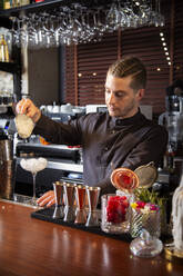 Konzentrierter junger männlicher Barkeeper in schwarzer Uniform gießt zerstoßenes Eis aus einem Shaker in ein elegantes Glas, während er einen erfrischenden Cocktail in einer Bar zubereitet - ADSF35252