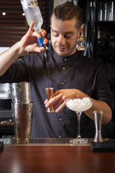 Positiver junger männlicher Barkeeper mit trendiger Frisur in Uniform gießt Wodka in einen Jigger, während er am Tresen steht und einen alkoholischen Cocktail mit Eis in einer Bar zubereitet - ADSF35253