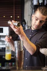 Konzentrierter junger professioneller Barmann in Uniform gießt Orangensaft aus einem Jigger in einen Shaker, während er einen erfrischenden Cocktail zubereitet und am Tresen einer Bar steht - ADSF35254