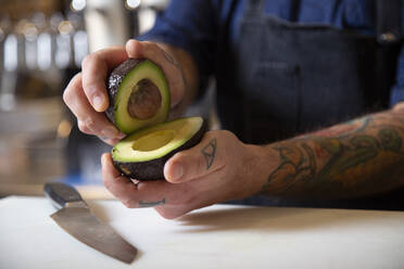 Cropped unrecognizable focused male chef in apron cutting ripe green avocado at table with cutting board while working in kitchen - ADSF35264