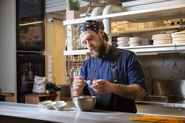 Fokussierter tätowierter männlicher Koch in Uniform, der frische rote Zwiebeln in eine Schüssel gibt, während er am Tisch in der hellen Küche eines Restaurants arbeitet - ADSF35266