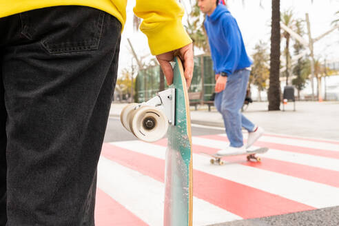 Schnittansicht von zwei gesichtslosen Skater-Freunden in blauen und gelben Pullovern, die an einem Zebrastreifen skaten - ADSF35269