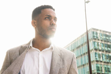 Nachdenklicher afroamerikanischer männlicher Unternehmer im eleganten Anzug schaut weg, während er in der Nähe eines modernen Bürogebäudes an einem sonnigen Tag in der Stadt steht - ADSF35282