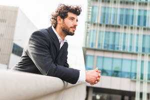 Side view of confident bearded male entrepreneur in elegant suit looking into distance while standing near contemporary high rise building on sunny street - ADSF35283