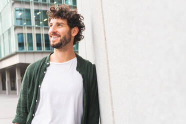 Thoughtful happy bearded male entrepreneur with black curly hair standing near wall of building on street of city - ADSF35287