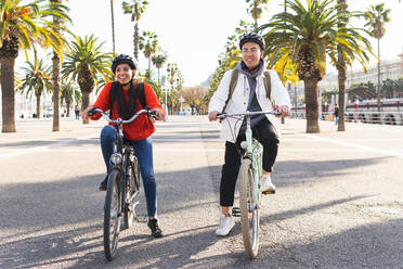 Full body of positive couple in helmets riding bicycles on asphalt road in city with green tress on summer day - ADSF35290