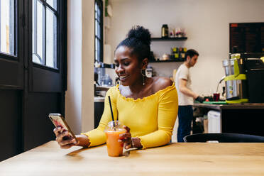 Attractive smiling African American female in bright yellow blouse holding disposable cup of fruit juice and browsing internet on smartphone while sitting at table in cafe - ADSF35327