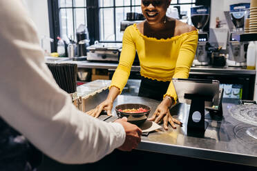 Glückliche afroamerikanische Kellnerin, die ein Tablett mit Smoothie-Schale und Löffeln einem unerkennbaren Kunden über den Tresen eines kleinen Cafés reicht - ADSF35328