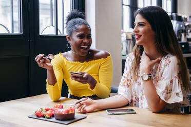 Young African American female reading message on smartphone while sitting together with friend in cafe and eating berry smoothie topped with strawberries - ADSF35332
