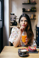Schöne fröhliche moderne Frau mit stilvollen Ohrringen genießt Fruchtsaft beim Frühstück sitzen am Holztisch im Cafe - ADSF35333