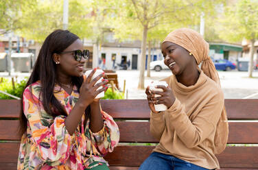 Positive junge afroamerikanische beste Freundinnen in legerer Kleidung, die sich lächelnd ansehen, während sie Kaffee zum Mitnehmen auf einer Bank im Stadtpark trinken - ADSF35350