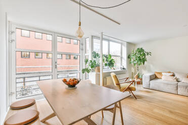 Esstisch aus Holz mit frischen Früchten in der Nähe von Glastüren in einer hellen, geräumigen Wohnung mit einem bequemen Sofa neben grünen Topfpflanzen am Fenster - ADSF35352