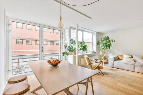Esstisch aus Holz mit frischen Früchten in der Nähe von Glastüren in einer hellen, geräumigen Wohnung mit einem bequemen Sofa neben grünen Topfpflanzen am Fenster - ADSF35352