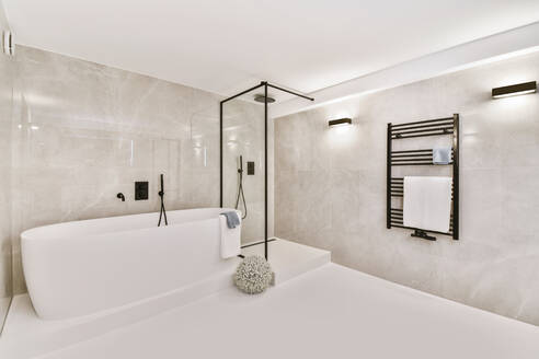 Weiße Keramikbadewanne neben einer Glasduschkabine in einem stilvollen geräumigen Badezimmer mit beheiztem Handtuchhalter und hell leuchtenden Lampen - ADSF35364