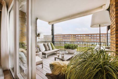 Comfortable sofas with cushions and armchairs placed near wooden table on terrace with glass entrance door of modern residential building - ADSF35382