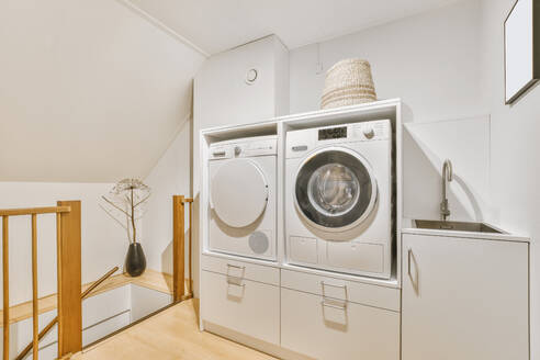 Moderne elektrische weiße Waschmaschine mit Trockner in der Nähe von Schrank mit Waschbecken in hellen Waschküche mit Holzzaun zu Hause platziert - ADSF35387