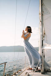 Trendy young female tourist in stylish outfit standing on deck of modern yacht sailing in sea and looking away during summer holidays - ADSF35388