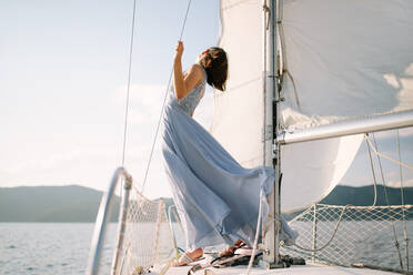 Trendy young female tourist in stylish outfit standing on deck of modern yacht sailing in sea and looking away during summer holidays - ADSF35389