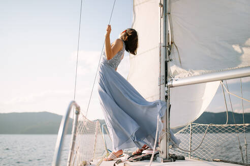 Trendy young female tourist in stylish outfit standing on deck of modern yacht sailing in sea and looking away during summer holidays - ADSF35389