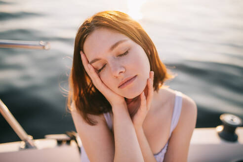 Ruhige junge Touristin mit braunen Haaren, die mit geschlossenen Augen ihr Gesicht berührt, während sie sich auf einer Yacht während einer Kreuzfahrt bei Sonnenuntergang entspannt - ADSF35391