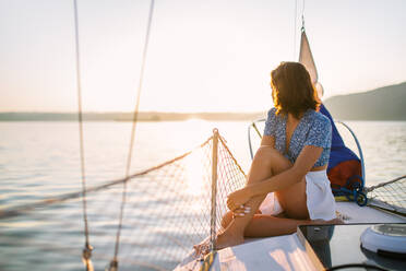 Seitenansicht von jungen stilvollen weiblichen Reisenden mit dunklen Haaren in trendigen crop top und Rock bewundern Meer beim Sitzen auf Segelboot während Kreuzfahrt unter Sonnenuntergang Himmel - ADSF35392