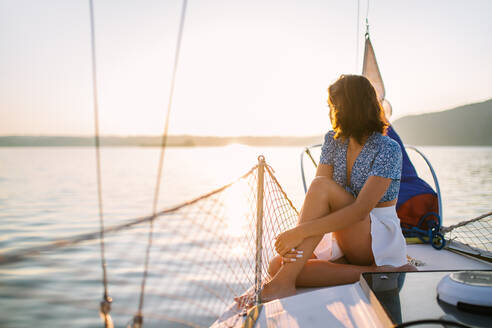 Side view of young stylish female traveler with dark hair in trendy crop top and skirt admiring sea while sitting on sailboat during cruise under sunset sky - ADSF35392