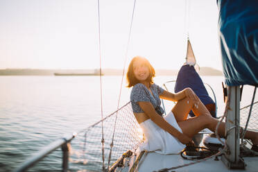 Side view of positive young stylish female traveler with dark hair in trendy crop top and skirt looking away while sitting on sailboat during cruise under sunset sky - ADSF35393