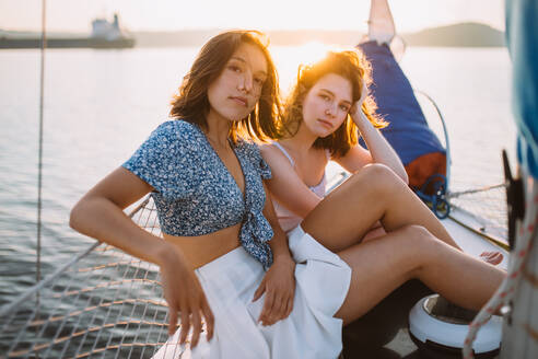 Seitenansicht von ernsthaften jungen Freundinnen in stilvollen Outfits entspannen auf Segelboot gegen Sonnenuntergang Himmel während des Sommerurlaubs - ADSF35394