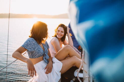 Side view of cheerful young female friends in stylish outfits smiling and talking while relaxing on sailboat against sunset sky during summer vacation - ADSF35395