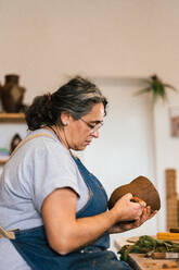 Ältere weibliche Keramikerin in Schürze benutzt ein Werkzeug und erstellt ein Muster auf einem Tontopf in einer Werkstatt - ADSF35397