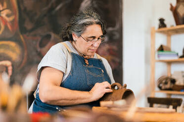Reife fokussierte weibliche Handwerkerin Keramikerin in Schürze Formung Stück Ton Topf in der Hand in der Werkstatt Tisch - ADSF35398
