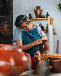 Reife fokussierte weibliche Handwerkerin Keramikerin in Schürze Formung Stück Ton Topf in der Hand in der Werkstatt Tisch - ADSF35399