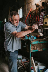 Fokussierter reifer männlicher Holzarbeiter, der eine Abrichtmaschine zum Hobeln von Holzbrettern benutzt, während er an einer schäbigen Werkbank in einer Tischlerwerkstatt arbeitet - ADSF35409