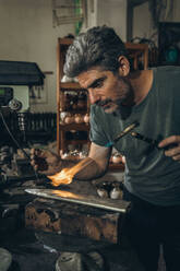 Bärtiger Handwerker mittleren Alters, der eine Gasfackel benutzt, um Details zu brennen, während er in der Nähe einer schäbigen Werkbank steht und tagsüber in der Werkstatt arbeitet - ADSF35424