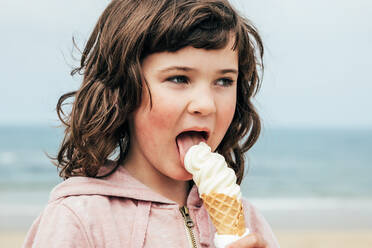 Mädchen mit dunklen Haaren schaut weg und leckt Eis, während sie Zeit auf unscharfen Hintergrund von Meer und Strand im Sommer verbringen - ADSF35425