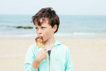 Süßer Junge mit grauen Augen, der wegschaut und süßes Schokoladeneis leckt, steht am Strand an einem Sommerwochenende - ADSF35426