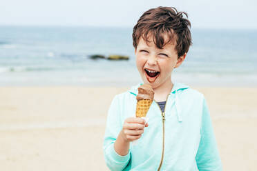 Netter fröhlicher Junge mit grauen Augen leckt süßes Schokoladeneis auf unscharfem Hintergrund von Strand am Sommerwochenende Tag mit geschlossenen Augen - ADSF35427