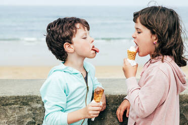Lustiger Junge mit schmutzigem Gesicht, der seiner Schwester die Zunge zeigt und Eis leckt, während er sich an der Grenze in der Nähe des wogenden Meeres an einem Wochenendtag am Strand abstützt - ADSF35428