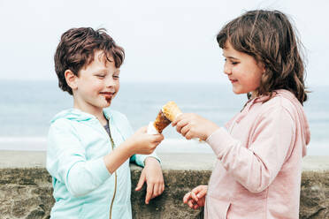 Cute brother and sister in casual clothes touching ice creams while standing near border on summer day on beach - ADSF35429