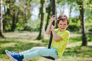 Seitenansicht eines entzückenden aufgeregten kleinen Jungen in Freizeitkleidung, der lächelt und in die Kamera schaut, während er auf einem Schaukelseil an einem sonnigen Tag im Park sitzt - ADSF35433