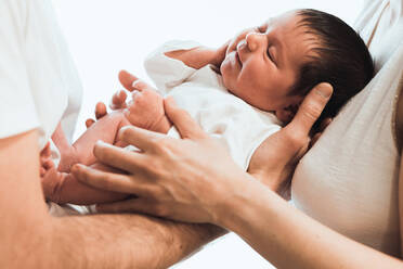 Crop unrecognizable young parent in casual clothes embracing adorable newborn baby with dark hair and closed eyes against white background - ADSF35439