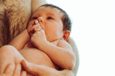 Adorable newborn baby with dark hair sucking hands while being embraced by crop unrecognizable shirtless father against white background - ADSF35441