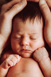 Von oben der Ernte anonyme Eltern streicheln Kopf der adorable nackten Neugeborenen mit dunklen Haaren liegen auf dem Bett mit geballten Fäusten und geschlossenen Augen - ADSF35443
