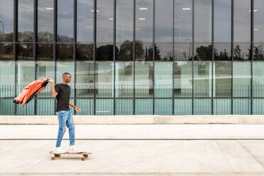 Seitenansicht eines trendigen jungen afroamerikanischen männlichen Millennials in Freizeitkleidung, der eine modische Jacke hält, während er in der Nähe eines modernen Gebäudes auf einer Straße Skateboard fährt - ADSF35449