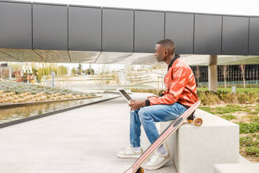 Seitenansicht eines konzentrierten jungen afroamerikanischen männlichen Freiberuflers in trendigem Outfit, der auf einer Steinbank sitzt und aus der Ferne an einem Laptop in der Nähe eines Longboards im Stadtpark arbeitet - ADSF35450