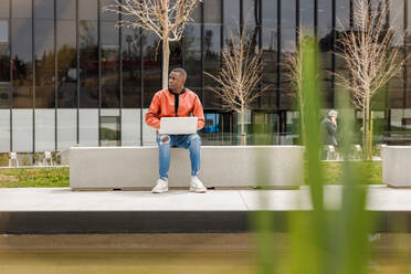 Fokussierter junger afroamerikanischer männlicher Freiberufler in trendigem Outfit, der auf einer Steinbank sitzt und aus der Ferne an einem Laptop im Universitätspark mit Teich arbeitet - ADSF35451