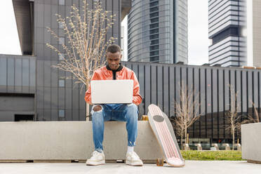 Konzentrierter junger afroamerikanischer Freiberufler in trendigem Outfit, der auf einer Steinbank sitzt und ferngesteuert an einem Laptop in der Nähe eines Longboards im Stadtpark arbeitet - ADSF35454